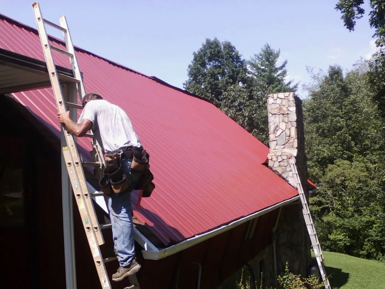Roofer on a ladder beside a red metal roof and stone chimney in Farmington